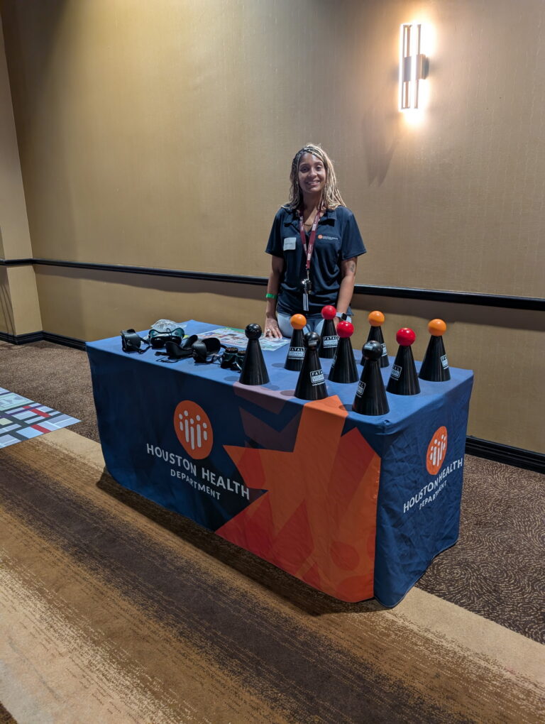 Shannon Starkey of the Houston Health Department stands behind a table displaying Fatal Vision impairment goggles and cone activities at a community outreach event