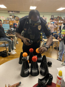 Police officer wearing Fatal Vision goggles reaches for colored balls balanced on Fatal Vision cones during a classroom activity