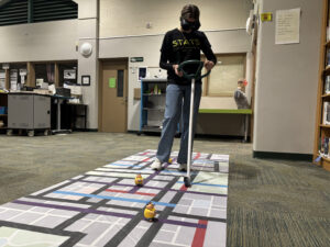 Student wearing Fatal Vision impairment goggles pushes a steering-wheel walker across a street-map floor mat with obstacle figures